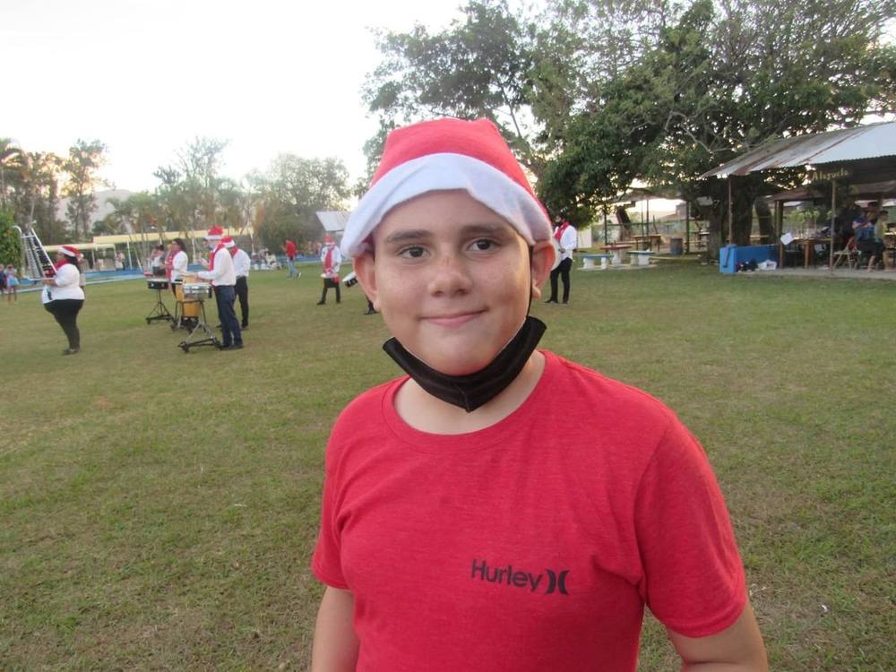 Un participante sonriente con gorro navideño; al fondo, una banda de percusión en un parque