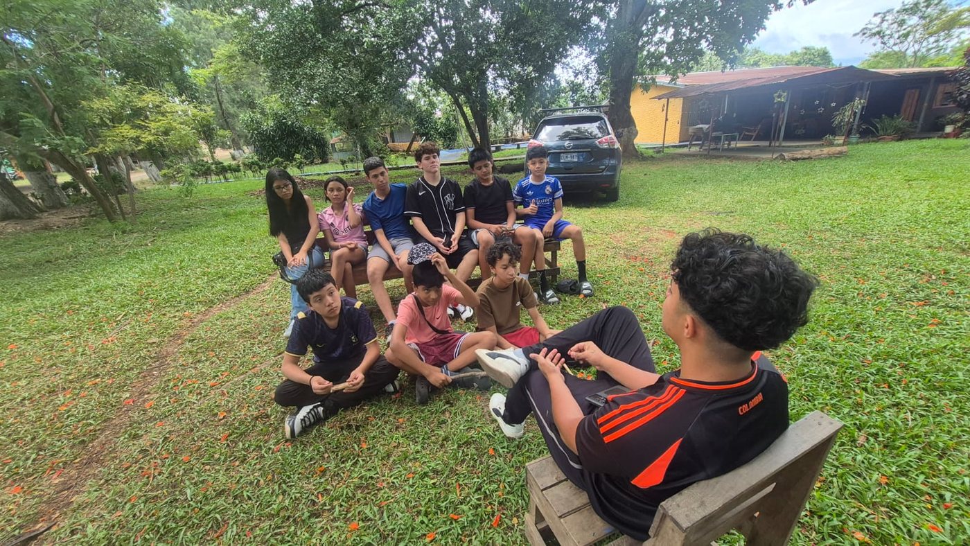 Grupo de participantes sentados al aire libre, entre árboles, escuchando a una persona facilitadora