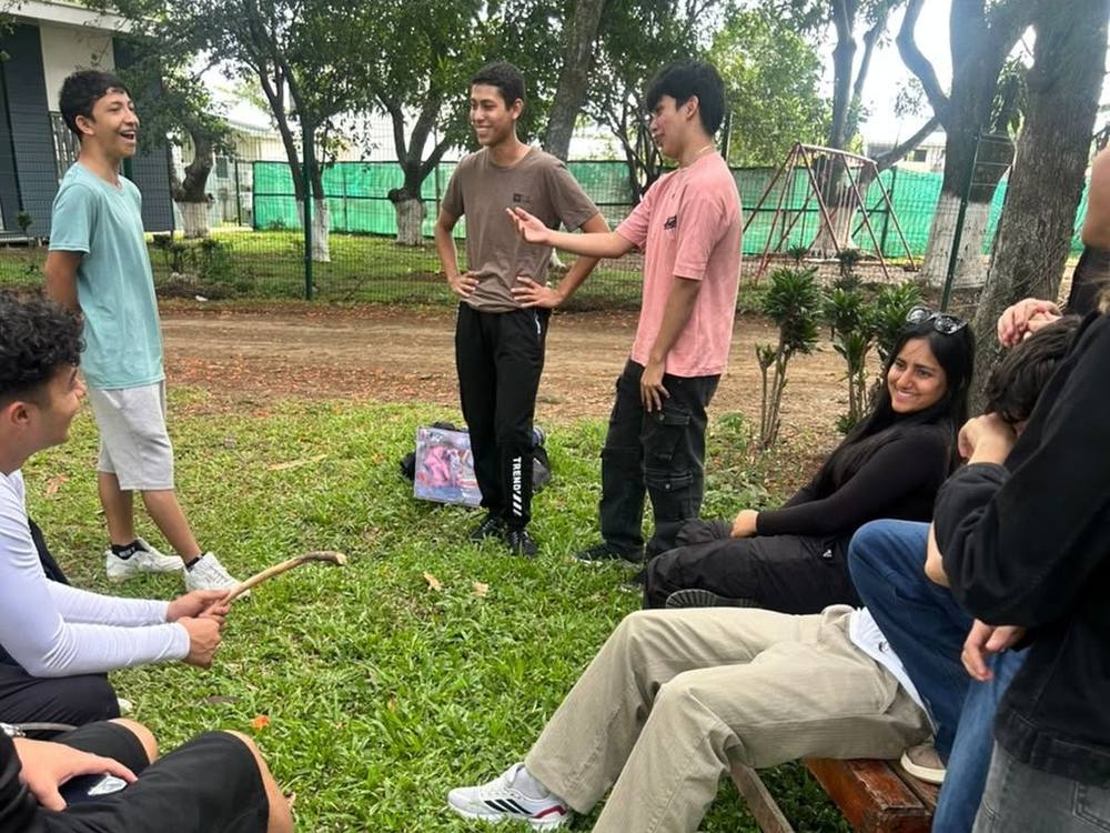 Jóvenes conversando y riendo al aire libre; unos de pie y otros sentados en bancas de madera