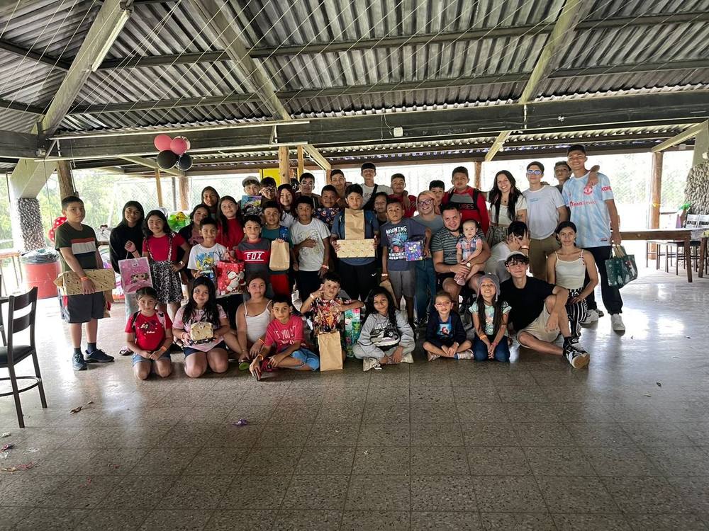 Foto grupal numerosa bajo un techo de lámina; niñas, niños, jóvenes y personas adultas sostienen regalos envueltos