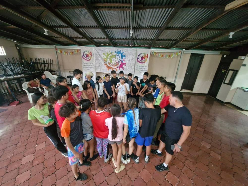 Participantes de pie formando un círculo en un salón, tomados de una cuerda, mientras una persona facilitadora habla al centro
