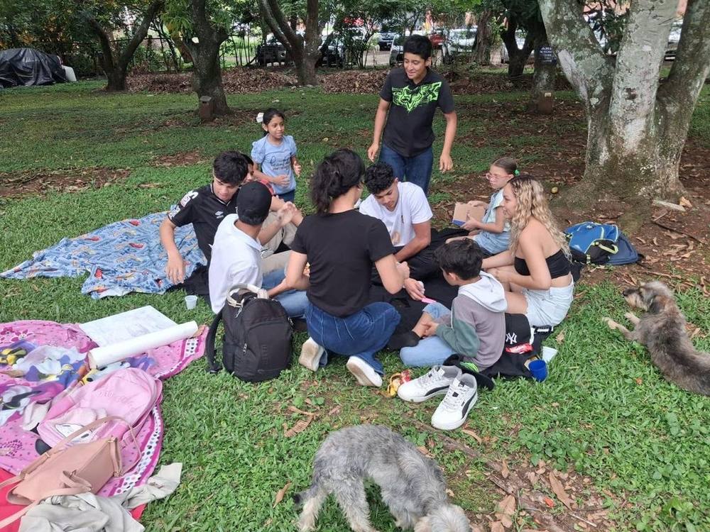 Un pequeño grupo sentado sobre una manta en el césped, bajo los árboles, compartiendo una actividad