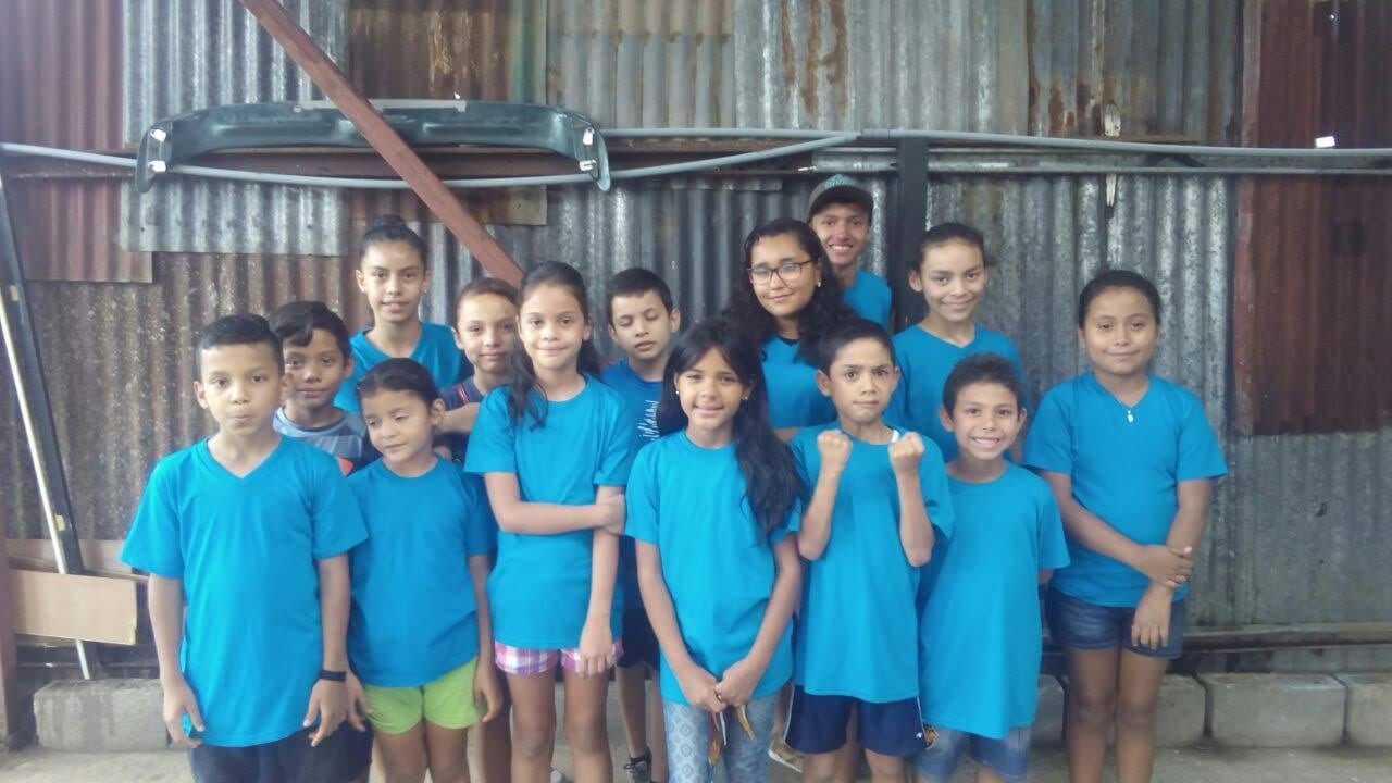 Grupo de niñas y niños con camisetas azul turquesa, posando juntos frente a una pared de lámina