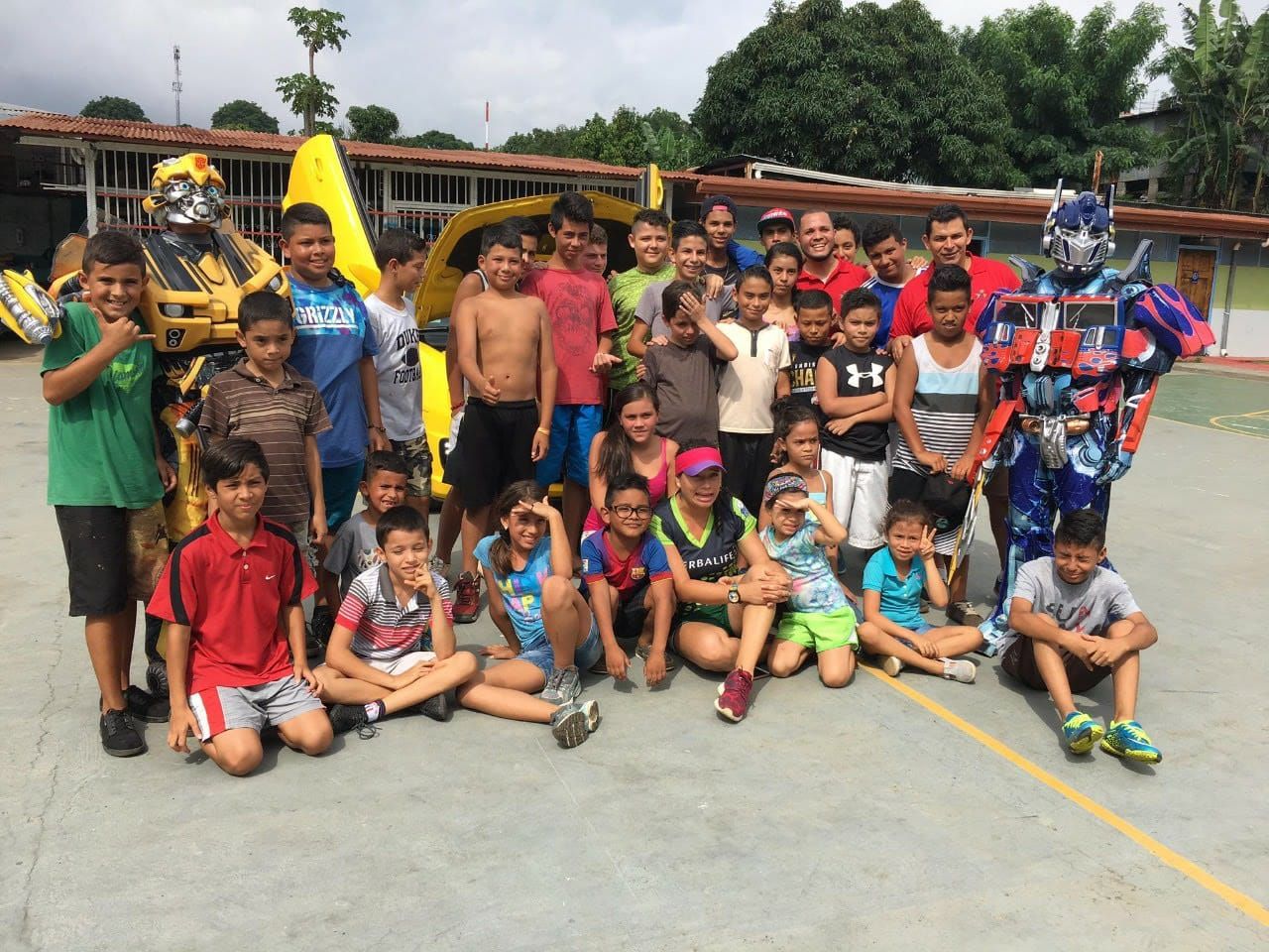 Foto grupal numerosa de niñas, niños y jóvenes en una cancha techada, junto a dos personajes disfrazados de robots