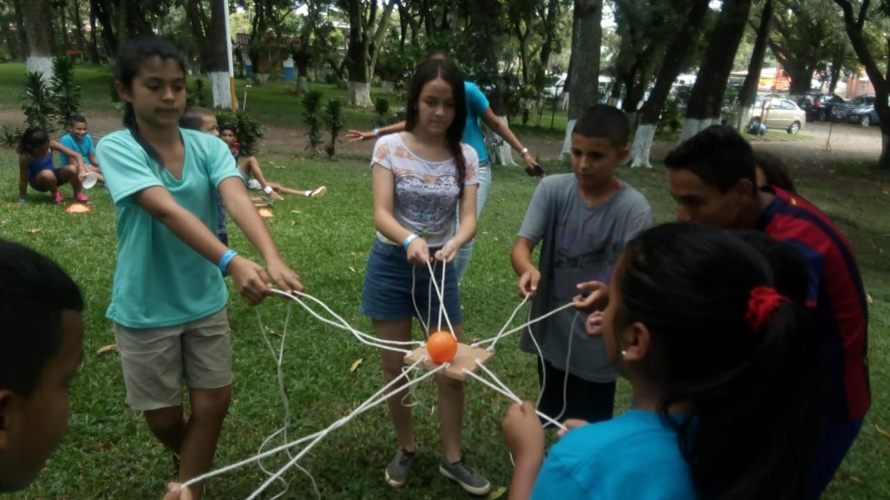 Un grupo de participantes sujeta cuerdas atadas a una pelota naranja, en un juego de coordinación sobre el césped