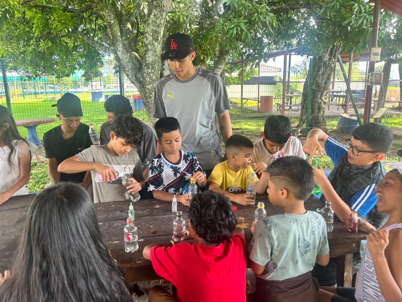 Niñas, niños y jóvenes sentados alrededor de una mesa de madera, al aire libre, trabajando con botellas en una actividad de taller; una persona joven de pie los acompaña