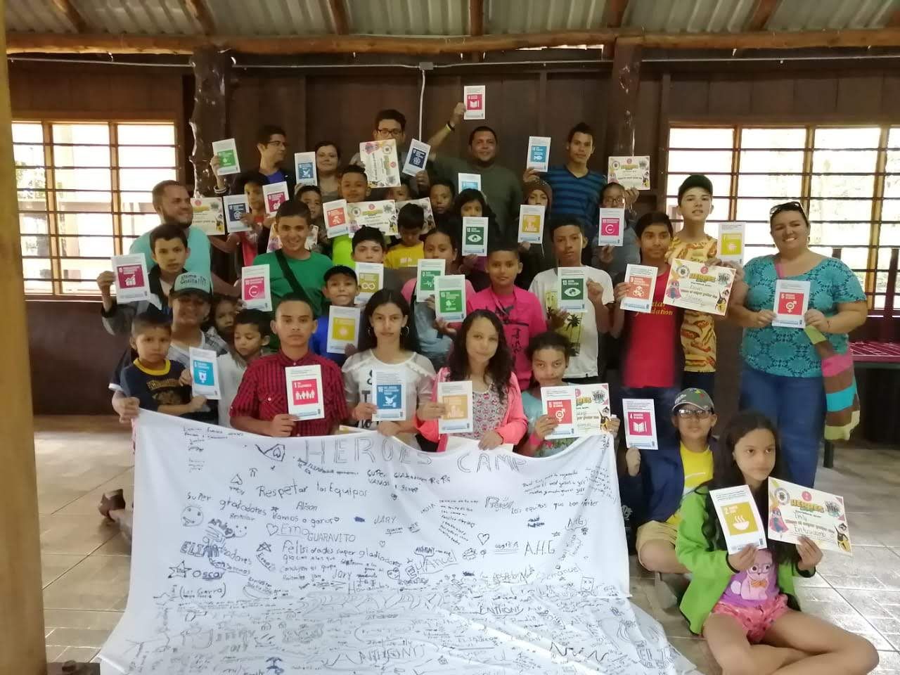 Grupo numeroso en un salón de madera; muchas personas sostienen tarjetas de colores con íconos y, al frente, despliegan una manta con mensajes escritos
